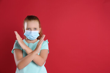 Little girl in protective mask showing stop gesture on red background, space for text. Prevent spreading of coronavirus