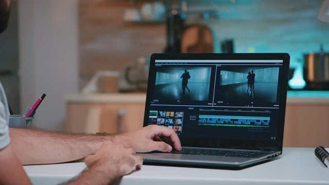 Videographer working on professional laptop from home, editing video and audio footage at night. Employee processing film montage on professional laptop sitting on desk in modern kitchen in midnight