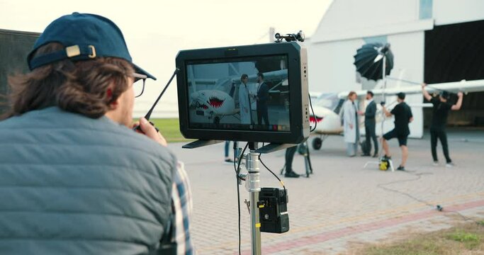 Rear of Caucasian male film director in cap looking at screen on movie set outdoor. Close up of filming equipment display. Males and females on background shooting scene near airplane. Filming concept