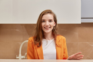 A young girl looks at the camera and smiles a snow-white smile. Bright orange jacket. In the background there is a kitchen and a water tap.