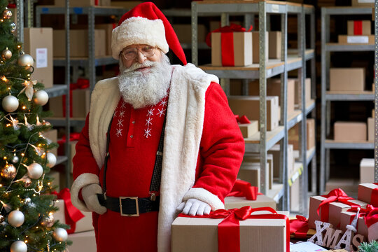 Happy old bearded Santa Claus wearing costume standing in workshop warehouse interior near Merry Christmas present gift box looking at camera. Xmas delivery concept. Portrait.