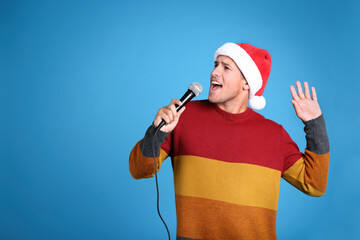Emotional man in Santa Claus hat singing with microphone on blue background, space for text....