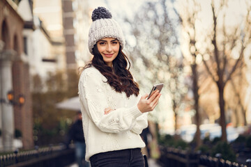 Elegant girl walk in a winter city. Woman in a white knited sweater. Beautiful lady with dark hair. Girl use the phone