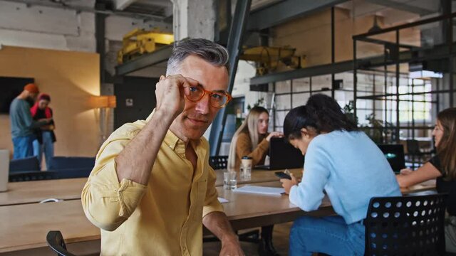 Mature man team leader sitting at desk, putting on glasses and smiling to camera in office, slow motion