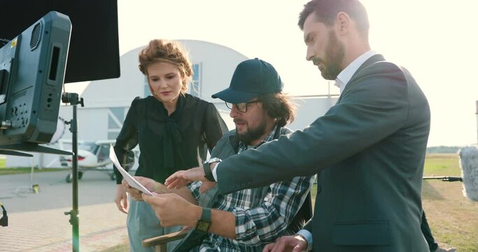Portrait of film crew members speaking outdoors on set. Handsome Caucasian male and beautiful female chatting with movie director on backstage. Filmmaker discussing script with actors. Filming concept