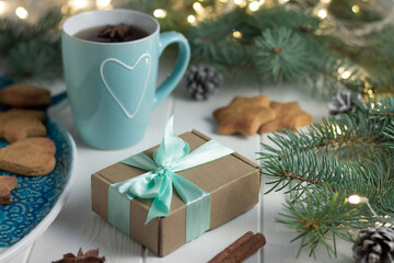 Fototapeta na wymiar Gift with mint ribbon, cup of tea, gingerbread, next to branches of Christmas tree with light bulbs on white background