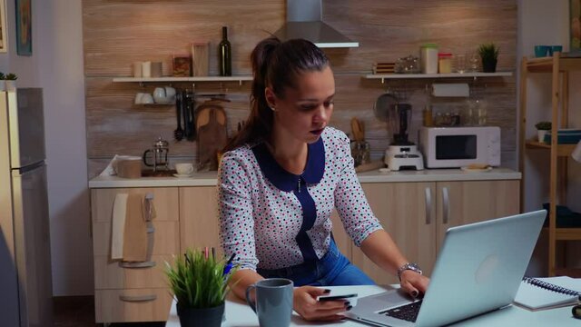 Hispanic lady holding credit card while doing online transaction using laptop in home kitchen late at night. Freelancer shopping online with electronic paymant on digital notebook conected to internet