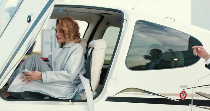 Handsome Caucasian man standing outdoor and watching beautiful young joyful red-haired female in coat getting into small airplane. Classy male actor on set with pretty woman. Movie scene concept