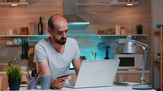 Freelancer making transaction from home using credit card sitting in the kitchen late at night. Freelancer shopping online from home with electronic paymant on digital notebook conected to internet