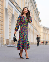 Obraz premium Elegant woman in dress walking city street on fall or autumn day. Pretty girl with long wavy hair holding handbag