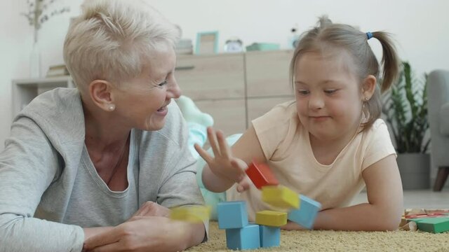 Medium shot of attractive elderly woman and her handicapped granddaughter lying on carpet in living room and playing with colorful tinker toys