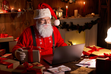 Happy old Santa Claus wearing hat holding gift box using laptop computer sitting at workshop home...