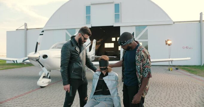 Mixed-races movie team on backstage outdoor with equipment. African American and Caucasian actors acting while making action film scene with gun and hostage.Film director with script talking to actors