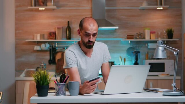 Young man shopping online late at night sitting in the kitchen on laptop using credit card. Employee making paymant transaction from home on digital notebook conected to internet