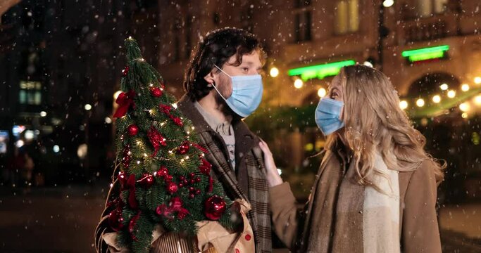Portrait of Caucasian happy family in masks standing outdoor and talking while snowing. Joyful man with beautiful woman on street with little new year tree. Close up. Christmas lights. Holiday concept