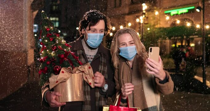 Close up portrait of Caucasian joyful wife and husband in masks outdoor and making selfie photo while snowing. Happy man with woman recording video on smartphone with new year tree. Christmas concept