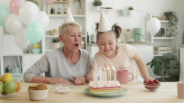 Medium shot of joyful aged woman and her granddaughter with down syndrome sitting at kitchen table and blowing out candles on birthday cake