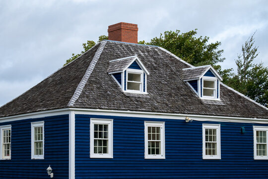 A vintage blue wooden building with white trim, double hung windows,  a wooden shingled roof, and a brown brick chimney. The old home has multiple stories with a green tree and grey skies. - Powered by Adobe