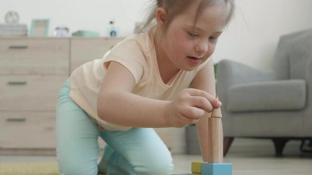 Medium shot of Caucasian handicapped girl with down syndrome sitting on carpet in living room and playing with colorful tinker toys
