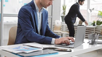 Medium of cropped Caucasian male office worker sitting at desk in office, typing on laptop, taking paper from folder and reading, colleague standing up and walking away in background