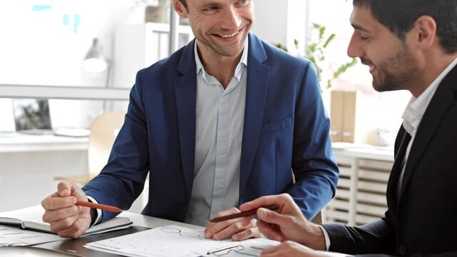 Over-the-shoulder of cropped male employee talking to middle-aged Caucasian business partner, discussing corporate project, looking at documents and laughing