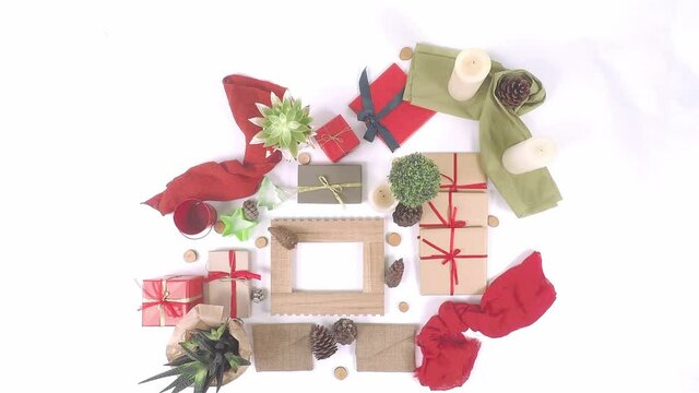 Top-down view of Christmas decorative items on white background with gift boxes, red ribbons, small candles, pine cones and small plants. Woman’s hand placing the items and stop motion animation.