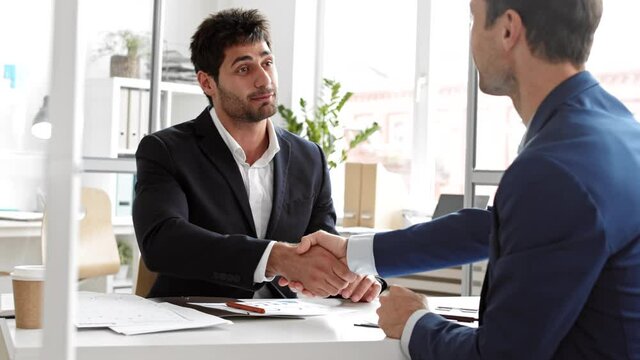Over-the-shoulder of unrecognizable man in blue suit sitting in workspace across desk from Mixed-Race young manager talking and laughing, shaking hands saying goodbye after successful job interview