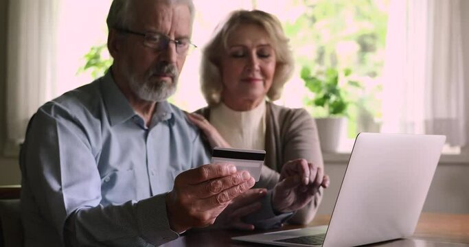 Smiling elderly couple sit at table using laptop and credit card make easy purchase on internet, shopping, paying on-line remotely. Older generation modern technology convenient comfort usage concept