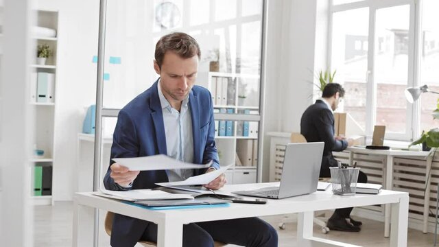 Medium tracking of male Caucasian business person dressed formally sitting at desk with laptop, smartphone in office, looking through documents then typing on keyboard 