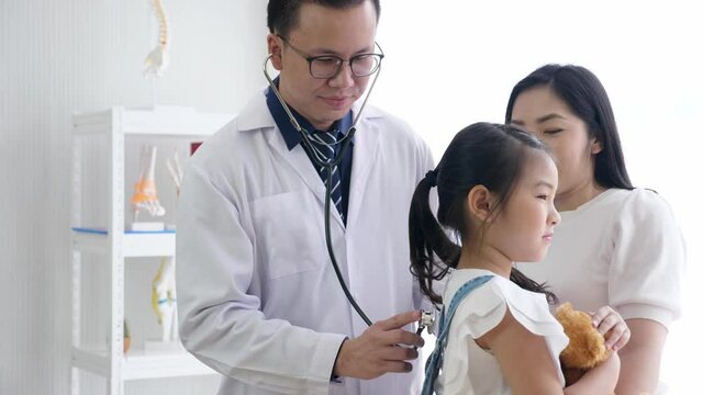 Asian pediatrician and kid concept. Middle aged male doctor puts stethoscope on the back of cute worried female child patient who is holding teddy bear with the mother standing besides.