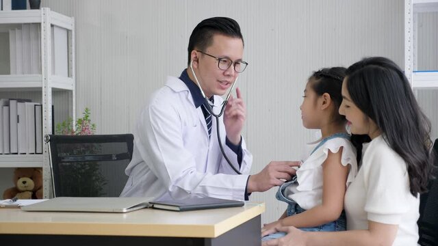Asian pediatrician and kid concept. Handheld, medium shot & pan shot of middle aged male doctor putting stethoscope in ears and listening to the chest of a cute girl who is sitting on mother's lap.