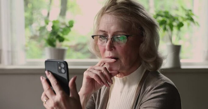 Close up aged woman in glasses sit indoor holding smart phone feels irritated. Older generation experiences problems, need help and explanation, lack of understanding with modern device usage concept
