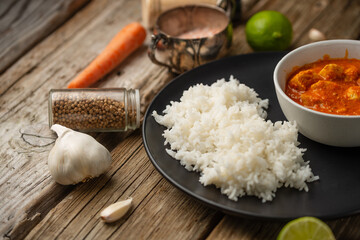 Black round plate with boiled rice and hot tasty chicken curry on rustic wooden table with lime and spices background. Classic Indian cuisine. Concept of tasty exotic spicy food.