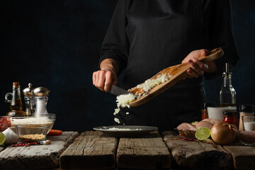 chef in black uniform pours chopped onion from wooden board into plate on rustic table with ingredients background. Frozen motion. Concept of cooking fresh healthy food with vegetables.