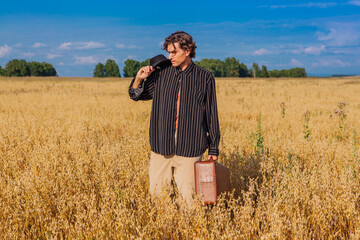 Naklejka na ściany i meble Tall handsome man with black hat and brown suitcase standing at golden oat field.