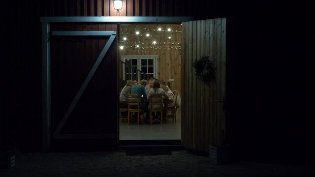 A Group Of People Sitting Around The Table Under The Garland Lights Seen From An Open Door At Night In Arendel, Zagorow, Poland. - wide shot