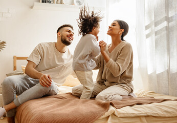happy diverse family mom, dad and child  laughing, playing and jumping   in bed   at home. - Powered by Adobe
