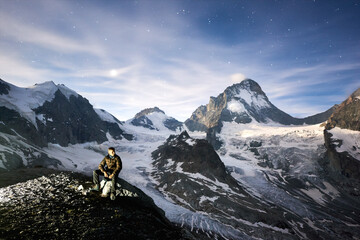 Amazing scenery in evening, man sitting on stone, beautiful mountains with white snow on background. Gorgeous mountain ridge with high rocky peaks, milky way with shining stars in sky, wonderland.