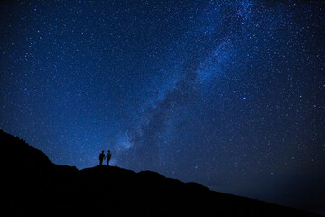 Naklejka na ściany i meble Silhouette of two girls / women on the hill. Stargazing at Oahu island, Hawaii. Starry night sky, Milky Way galaxy astrophotography.