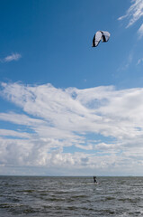 In the distance, unrecognizable kitesurfer girl in black suit, holding black and white kite, moves on board leaving streak of spray on bay of sea coast against blue sky densely covered with clouds.