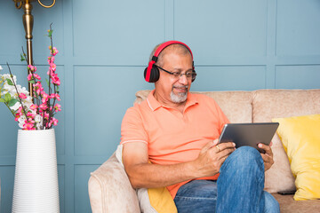 Indian retired senior adult man using headphones with smartphone or tablet for entertainment