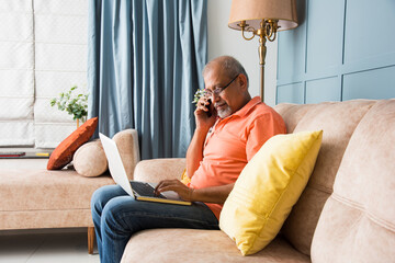 Indian asian old man speaking on phone while using laptop computer at home