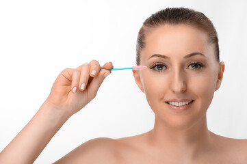 A beautiful light-skinned girl with beautiful eyes takes care of her extended eyelashes with an eyelash brush.  Soft light isolated white background.