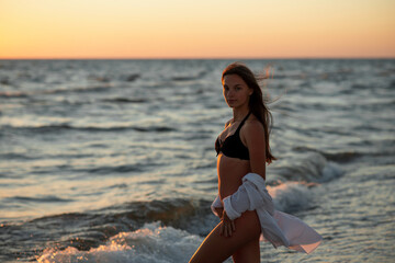 Fototapeta na wymiar Young beautiful woman in black bikini and white shirt on a tanned body on the beach at sunset. Soft selective focus, art nose.