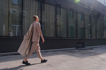 A woman with short hair in gray coat and glasses dancing, walking, smiling on a city street.