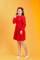 Cheerful young Asian woman in red dress talking on phone, standing over orange background. Full length. Full body.