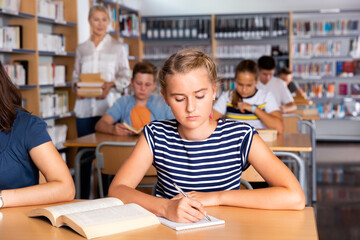 Fototapeta na wymiar Diligent serious schoolgirl writing lesson in classroom, teacher on background