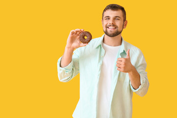 Handsome young man with sweet donut showing thumb-up on color background