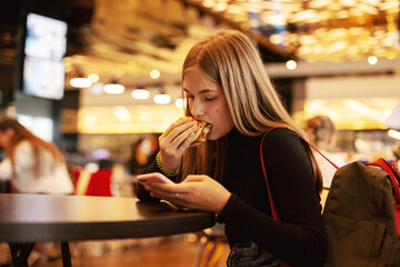 A beautiful teenage girl sitting in a cafe looks at the phone and eats a hamburger. Happy child eating junk food in a restaurant.