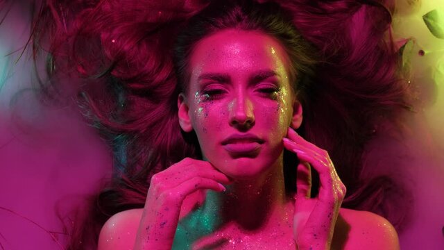 Portrait of a girl with glitter on her body in pink creative light. The girl lies in flowers and smoke. Creative make-up. The image of a nymph.
